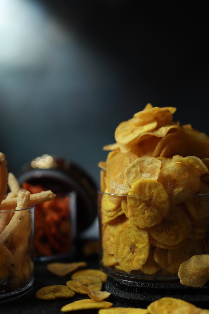 Close-up of assorted banana chips in glass jars with a moody background.